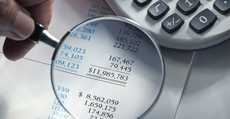 Hand holding magnifying glass over financial documents with large numbers, next to a calculator