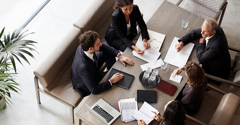 Five business professionals meeting around a table, discussing documents and working on laptops, to symbolize perspectives