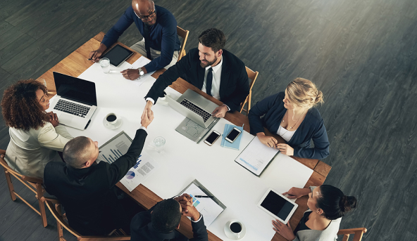 Business team meeting around a table with laptops and tablets, two men shaking hands