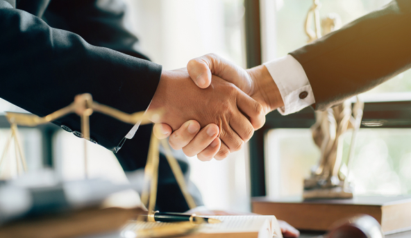 Two professionals in suits shaking hands in an office, with legal scales and Lady Justice statue in the background