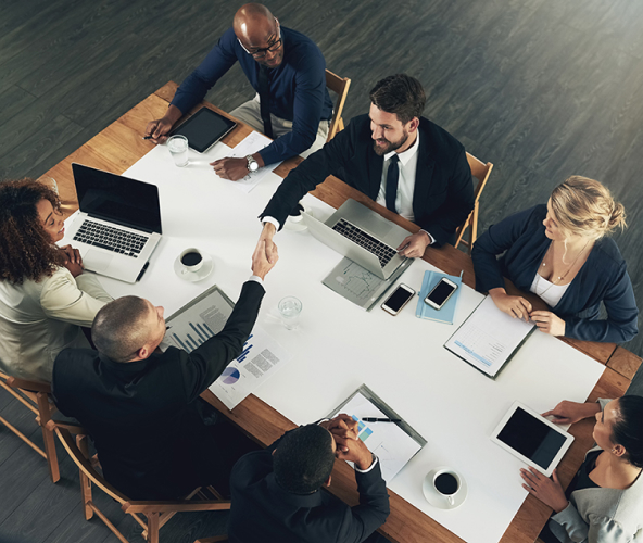 Business team meeting around a table with laptops and tablets, two men shaking hands