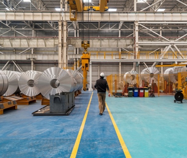 A man walks among large aluminum rolls in a factory.